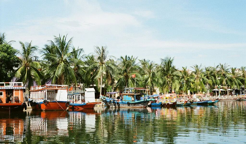 Bunte Boote am Flussufer von Hoi An, umgeben von Palmen und traditioneller Architektur